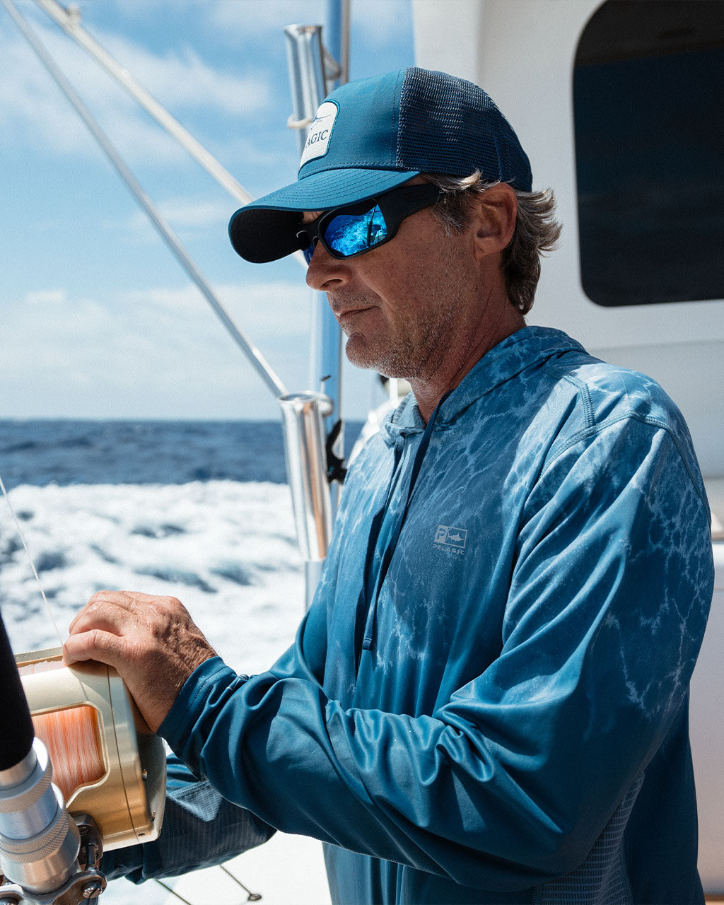 Man on a sailboat wearing a blue cap and sunglasses, holding a fishing reel.